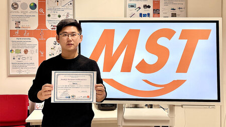 艾恩·荣格（Ein junger asiatischer Mann mit Brille hält Ein Urkunde）在哈登登。提示是Buchstaben MST die Abkürzung der Professsur Mess-und Sensortechnik zu sehen。