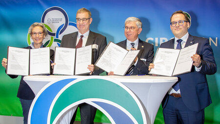 一名女子和三名男子站在一张高桌子后面，手里拿着签署的合同文件。