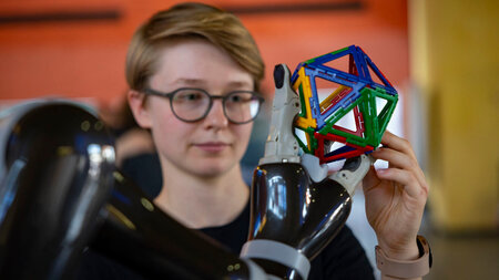 Eine junge Frau blickt auf Eine Roboterhand，die in buntes geometricsches Gebilde festhält.埃因·荣格夫人在机器人手上发呆，在几何图形中死去。