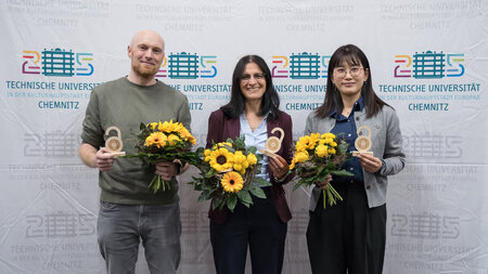 Ein Mann und zwei Frauen stehen nebeneinander. IN den Händen halten sie einen kleinen hölzernen Pokal und einen Blumenstrauß.