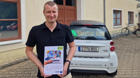 Ein Mann steht vor einem Auto und hält eine Urkunde in der Hand.