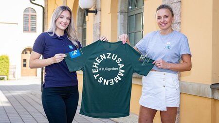 Zwei junge Frauen halten gemeinsam ein grünes T-Shirt hoch.