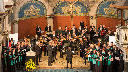 Mehrere Musikerinnen und Musiker sowie Sängerinnen und Sänger stehen im Altrraum einer Kirche.