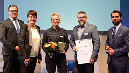 Drei Männer und zwei Frauen stehen nebeneinander. Eine hält einen Blumenstrauß und einer eine gerahmte Urkunde in den Händen.