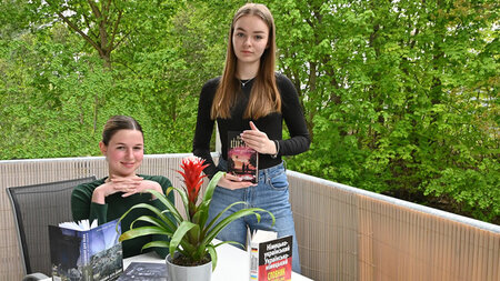 Zwei Mädchen sitzen bzw. stehen auf einem Balkon an einem Tisch, auf dem Bücher liegen.
