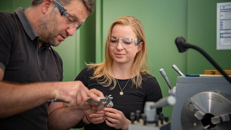 Ein Mann erklärt einer Frau ein technisches Bauteil und misst mit einem Messschieber den Durchmesser des Teils. Beide tragen eine Schutzbrille. 
