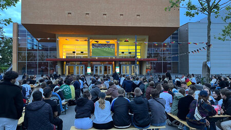 Mehrere Personen sitzen auf Bänken und blicken auf einen großen Bildschirm an einem Gebäude.