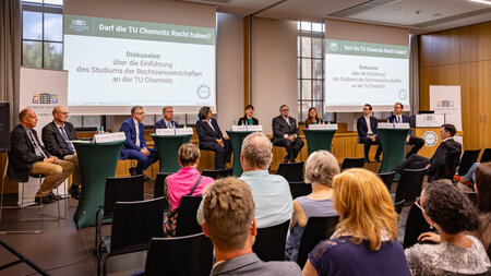 Mehrere Personen sitzen im Podium, weitere Personen sitzen im Raum und blicken nach vorn. 