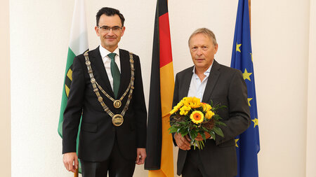 Zwei Männer im Anzug stehen nebeneinander, einer hält einen Blumenstrauß in den Händen, der andere trägt eine goldene Amtskette.