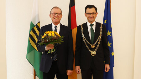 Zwei Männer im Anzug stehen nebeneinander, einer hält einen Blumenstrauß in den Händen, der andere trägt eine goldene Amtskette.