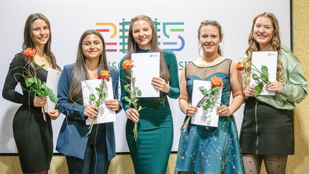 Junge Frauen stehen nebeneinander mit Blumen und Zeugnismappen in den Händen. 