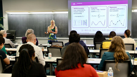 Eine FRau steht vor einer Tafel und spricht zu den Zuhörern und Zuhörerinnen in einem Seminarraum.