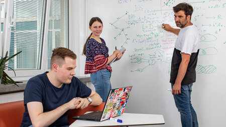 Ein Mann und eie Frau stehen an einer Tafel, ein Mann sitzt vor ihnen an eienm Tisch und arbeitet am Laptop.