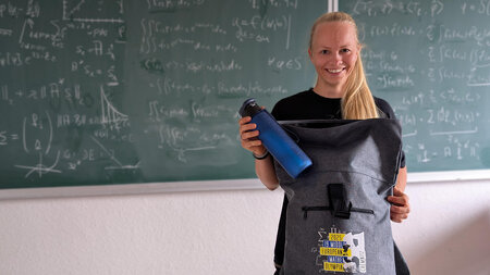 Eine Frau steht vor einer mit Formeln beschriebenen Tafel und packt eine Trinkflasche in einen Rucksack.