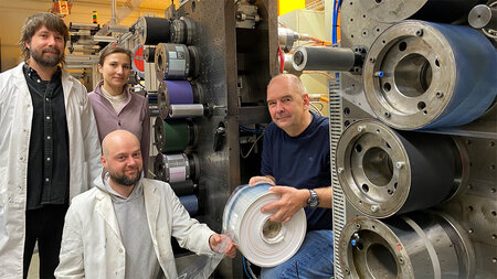 Drei Männer und eine Frau stehen bzw. kniene neben einer Druckmaschine, ein Mann hält eine Rolle in den Händen.