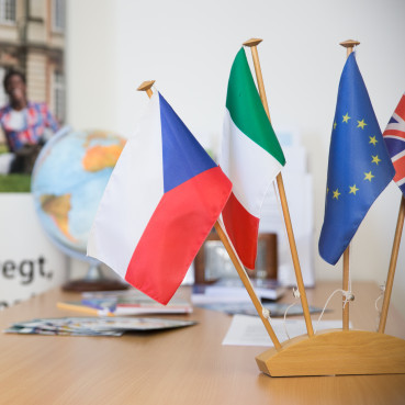 Flaggen verschiedener Länder auf einem Tisch im Vordergrund, Globus und Plakat über Erasmus-Programm im Hintergrund